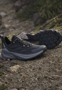 Chaussures de trail noires et grises avec semelles robustes posées sur un sol rocheux en plein air près de la verdure.
