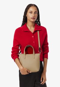 Borsa a mano in pelle marrone chiaro con finitura testurizzata, doppio manico e tracolla regolabile. Modello che indossa un maglione rosso a maglia e pantaloni neri.