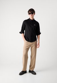 Zwarte overhemdd met een klein wit logo, beige broeken en zwarte schoenen; model met een donkere zonnebril, staand tegen een effen achtergrond.