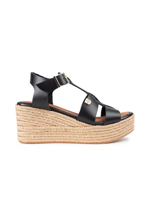 Sandal de cuña negra de cuero con suela de yute trenzada, correa ajustable en el tobillo y diseño entrecruzado. Acento de hardware dorado en la correa.