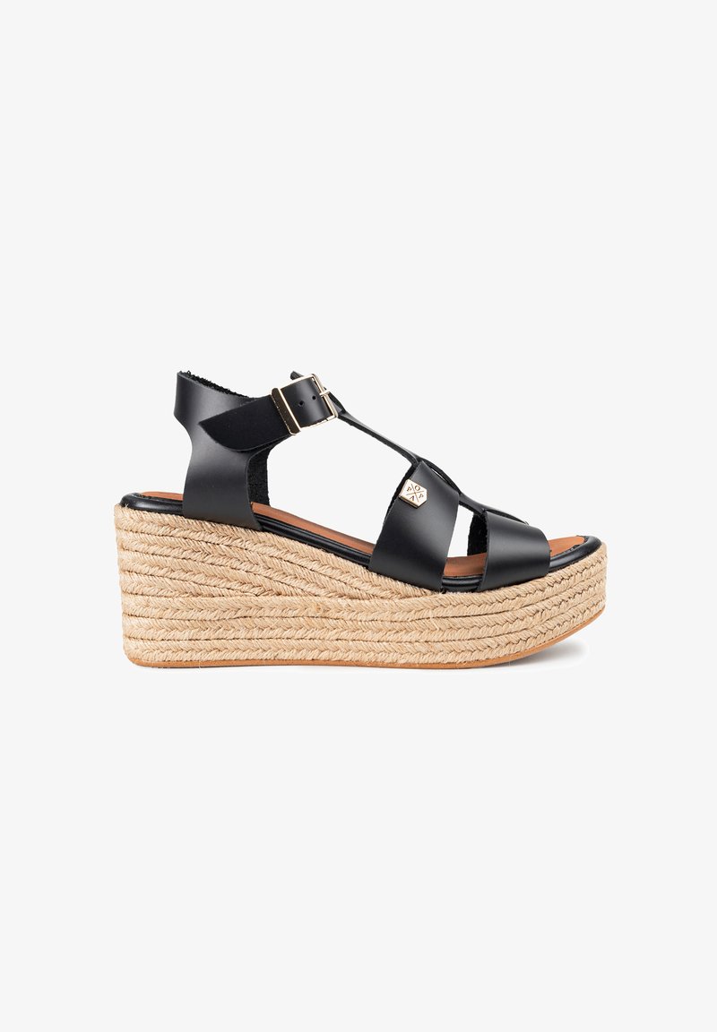 Sandal de cuña negra de cuero con suela de yute trenzada, correa ajustable en el tobillo y diseño entrecruzado. Acento de hardware dorado en la correa.