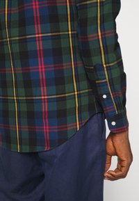 Koszula w kratkę w kolorach zielonym, granatowym, czerwonym i żółtym. Zapięcie na guziki przy mankietach i zaokrąglony dół. Wykonana z materiału bawełnianego o miękkiej teksturze.