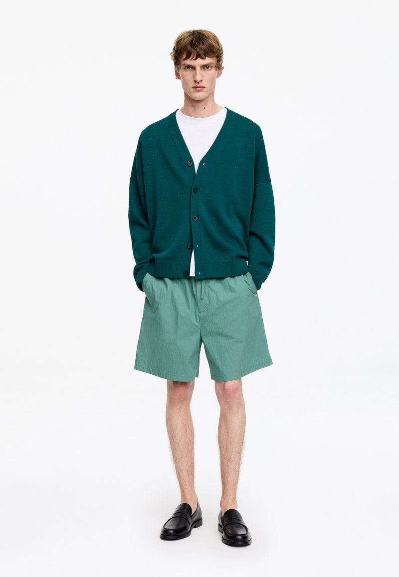 Jeune homme debout, les mains dans les poches, portant un cardigan vert foncé, un T-shirt blanc, un short vert clair et des mocassins noirs sur fond blanc.