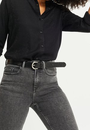 Kobieta w czarnej, teksturowanej koszuli zapinanej na guziki, włożonej do wysokich, wyblakłych czarnych dżinsów z czarnym skórzanym paskiem z srebrną klamrą.