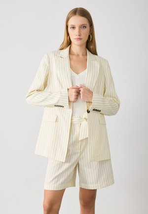 Pruhovaný dvoudílný outfit s off-white sakem s černými pruhy a odpovídajícími šortkami, vyrobený z lehkého materiálu s předními kapsami.