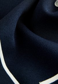 Námornícka modrá tkanina s textúrovaným povrchom, ktorú zdobí biely akcentový vzor, prelínajúci sa cez materiál v zakrivenom dizajne.
