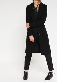 Femme portant un long manteau noir, un pantalon noir, des bottines noires et un haut vert foncé, debout, les bras croisés et les jambes écartées.