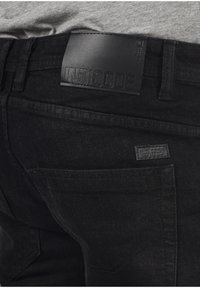 Jeans in denim nero con una toppa in pelle testurizzata etichettata "INDICODE" sulla vita e una piccola etichetta in tessuto sulla tasca posteriore.