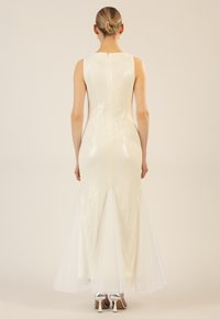 Un abito da sera bianco con silhouette aderente, caratterizzato da una gonna in tulle trasparente e una cerniera sul retro discreta. Visibili delle scarpe alte argentate.