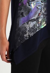 Hand som bär mörka byxor och en marinblå blus med lila påfågel- och blommönster, fjärilskonturer och asymmetrisk fåll.