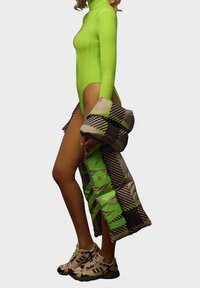 Neongrön ribbad bodysuit med hög krage, tillsammans med en vadderad, mönstrad kappa i beige, grön och svart. Beiga träningsskor.