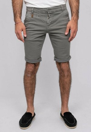 Homme portant un short gris retroussé, une chemise blanche, des chaussures noires sans lacets, debout les mains détendues le long du corps devant un fond uni.