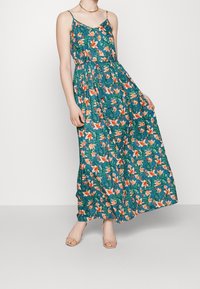 Kvinna som bär en turkos blommig ärmlös maxiklänning med tunna band och öppettåiga beige sandaler, stående mot en enkel vit bakgrund.