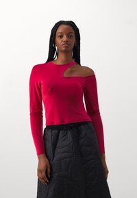 Fitted long-sleeve top in bright pink fabric, featuring a unique cutout design on one shoulder. Paired with a black quilted skirt.