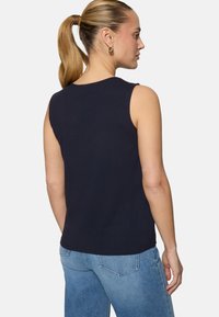 Femme avec queue de cheval blonde portant des boucles d'oreilles créoles dorées, un haut bleu marine sans manches et un jean bleu clair, vue de dos et de profil.