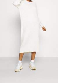 Abito lungo a maglia a coste color bianco sporco con scollatura a V e maniche lunghe. Abbinato a sneaker bianche con dettagli beige e calze bianche.