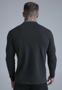 Muškarac s detaljnom tetovažom na vratu, obučen u crnu majicu s dugim rukavima, stoji okrenut leđima prema kameri na jednostavnoj sivoj pozadini.