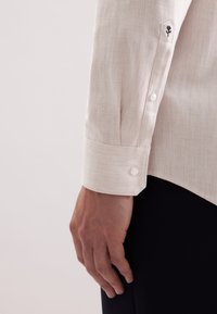 Beżowa koszula w jodełkę z mankietem na guzikiem, z małym czarnym kwiatowym haftem na rękawie. Gładka tekstura i dopasowany krój.