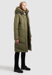 Abrigo de plumas verde oliva con cuello alto, patrón acolchado, botones en la parte frontal y bolsillos laterales, usado sobre pantalones ajustados negros y botas.