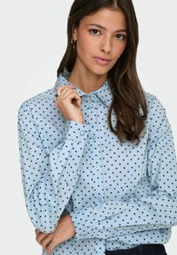 Chemise boutonnie bleu clair avec des pois navy, design col et manches longues. Le tissu semble doux avec une texture lisse.