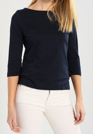 Kobieta ubrana w dopasowaną granatową bluzkę z rękawem do połowy przedramienia i jasnobeżowe spodnie z wysokim stanem, stojąca na jednolitym tle.