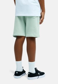 Παιδί που φοράει ανοιχτό πράσινο σορτς, λευκές κάλτσες και μαύρα αθλητικά παπούτσια της μάρκας DC, στέκεται με τα πόδια ορατά από το μέσο του μηρού και κάτω.