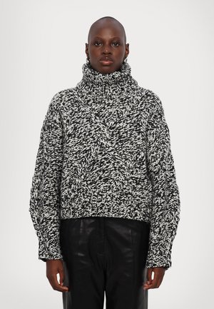 Suéter grueso de punto en blanco y negro con un cuello alto, patrón de trenzas y puños acanalados. Llevado con pantalones de cuero negros.