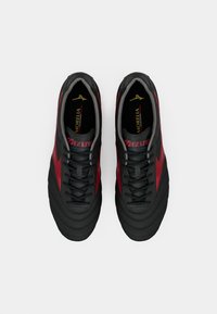 Chuteiras de futebol em preto e vermelho com parte superior sintética texturizada, atacadores planos e colarinho acolchoado; apresenta o logótipo e o nome da marca na língua.