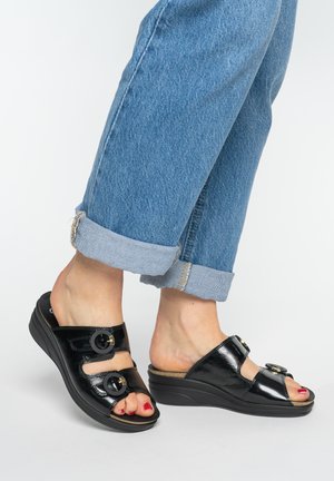 Pies con sandalias de cuña de charol negro con dos hebillas, combinadas con vaqueros azules con puños y esmalte de uñas de los pies rojo.