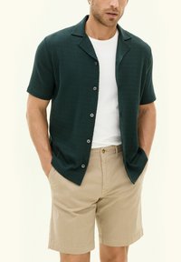 Tummanvihreä lyhythihainen paita, jossa on teksturoitu kuvio, napit edessä, yhdistettynä vaaleanruskeisiin shortseihin. Paidassa on kaulus ja harmaat napit.