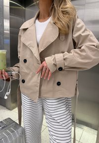 Giacca beige doppiopetto con grandi bottoni neri, ampio colletto e maniche lunghe, abbinata a pantaloni a righe. La persona tiene in mano una bevanda e cuffie.