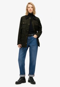 Chaqueta de utilitaria verde oscura con múltiples bolsillos, botones dorados, suéter negro de cuello alto, jeans de denim azul sueltos y botas negras con cordones.