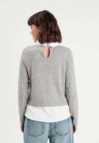 Grauer gestrickter Pullover mit langen Ärmeln, Rundhalsausschnitt und einem Schlüssellochdetail im Rücken, über einem weißen Hemd getragen. Besitzt eine strukturierte Oberfläche.