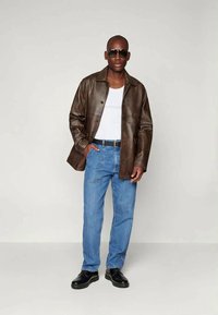 Rjava usnjena jakna s ovratnikom, bela brezrokavnik, modri kavbojke in črne čevlje. Model nosi temne sončne očala. Minimalna ozadje.