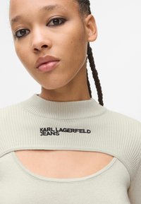 Šviesiai pilka raižyta megztinė su aukštu kaklu, su iškirpimo detalėmis ir juoda siuvinėta "Karl Lagerfeld Jeans" logotipu.