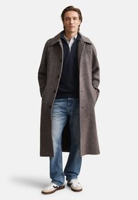 Garš pelēks pundurzīdu mētelis ar notiklu apkakli un pogu nosegumu, valkāts pār tumši zilu džemperi, komplektā ar zilām džinsām un baltām sporta kurpēm.