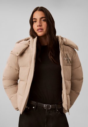 Ung kvinde med langt mørkt hår iført en beige Calvin Klein dunjakke over en sort skjorte og sorte bukser med bælte.