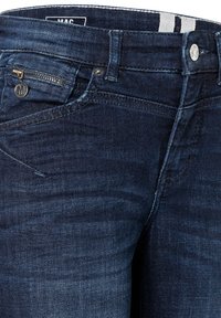 Vaqueros de mezclilla azul oscuro con un corte ajustado, que cuentan con un pequeño bolsillo con cremallera, botón de marca y sutiles detalles de desgaste en la tela.