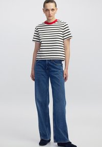 Mujer vestida con una camiseta a rayas blancas y negras con cuello rojo, jeans anchos de mezclilla azul y zapatos oscuros, de pie frente a un fondo liso.