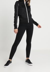 Sudadera negra con cremallera y detalles en blanco, combinada con jeans skinny negros y zapatillas negras. Tejido suave y diseño ajustado.