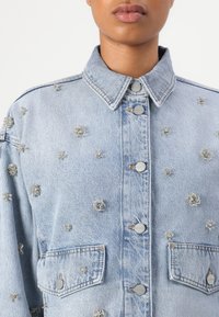 Nahaufnahme einer Person, die eine hellblaue Denimjacke mit silbernen Perlenverzierungen und geknöpftem Vorderteil trägt, Kragen sichtbar, kein Gesicht gezeigt.