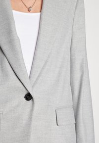 Blazer gris avec un revers cranté, fermeture à un bouton noir unique et deux poches avant. Le tissu a une texture lisse avec un motif subtil.