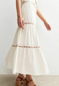 Jupe maxi blanche avec broderies florales en rose et vert, présentant un design en couches et un tissu fluide. Sandales à talons carrés et lanières marron.