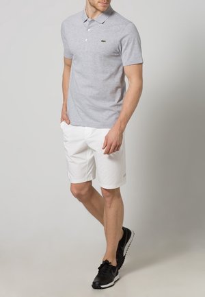 Homme portant un polo gris clair, un short blanc jusqu'aux genoux et des chaussures de sport noires, se tenant les mains dans les poches.