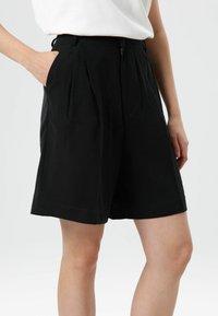 Zwarte shorts met hoge taille, voorplooien, zijzakken en een gladde textuur. Voorzien van een ritssluiting en ceintuurlussen.