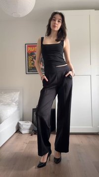 Femeie în picioare într-o cameră, purtând un top negru fără mâneci, pantaloni negri cu picioare largi și pantofi stiletto negri, cu mâinile în buzunare.