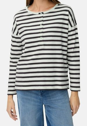 Frau trägt ein langärmeliges Oberteil mit schwarz-weißen horizontalen Streifen und vier Knöpfen an der Blende, kombiniert mit blauen Jeans, steht mit entspannten Armen.