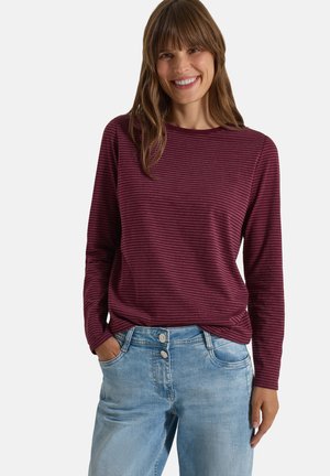 Femme aux cheveux bruns mi-longs souriante, portant une chemise rayée bordeaux à manches longues et un jean en denim bleu clair.
