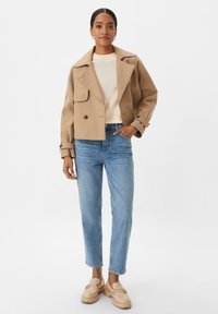 Cappotto trench cropped beige con ampi rever e chiusura a doppio bottone, abbinato a jeans in denim chiaro e scarpe slip-on beige.