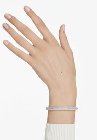 Bracelet en argent avec de délicates pierres transparentes alternées, présentant un design lisse et plat. Porté sur une main avec du vernis à ongles de couleur claire.